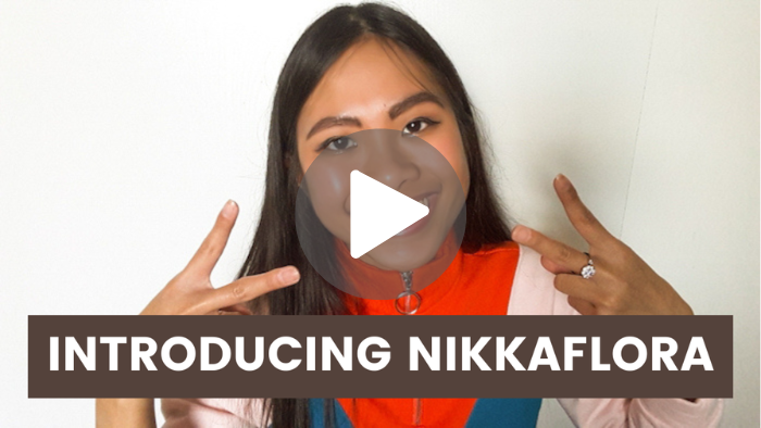In this photo, this is a youtube thumbnail of Nikkaflora’s first video, which is called “Introducing Nikkaflora.” Nicole from Nikkaflora is smiling widely at the camera while showing off two peace signs with her hands. She is wearing natural-toned eye makeup on her face, along with sporting an orange, blue, and light pink jacket. The words Introducing Nikkaflora are written in a bold font on top of a dark brown rectangle. Image by © Nikkaflora.